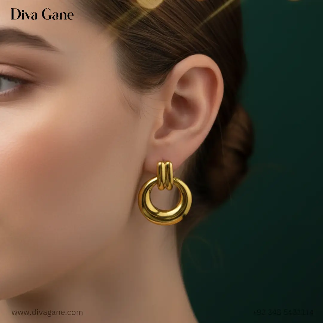 duality dots Close-up of a woman's ear wearing a bold, gold-finished earring. The earring features a thick, polished circular hoop suspended from a double-bar gold post. The background is a dark emerald green silk with warm, out-of-focus golden bokeh lights, creating a luxury aesthetic