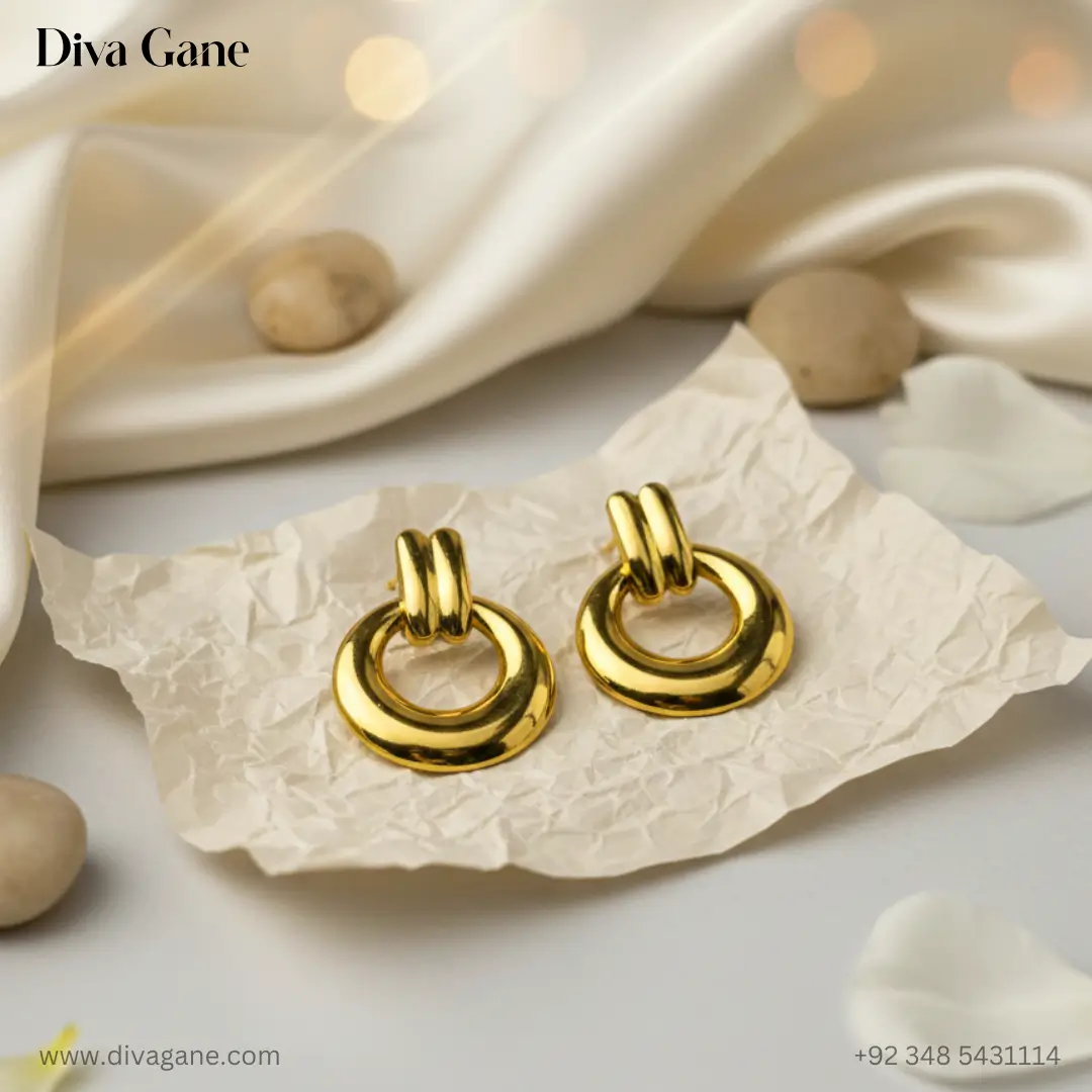 duality dots gold-finished earring. The earring features a thick, polished circular hoop suspended from a double-bar gold post. The background is a dark emerald green silk with warm, out-of-focus golden bokeh lights, creating a luxury aestheticots