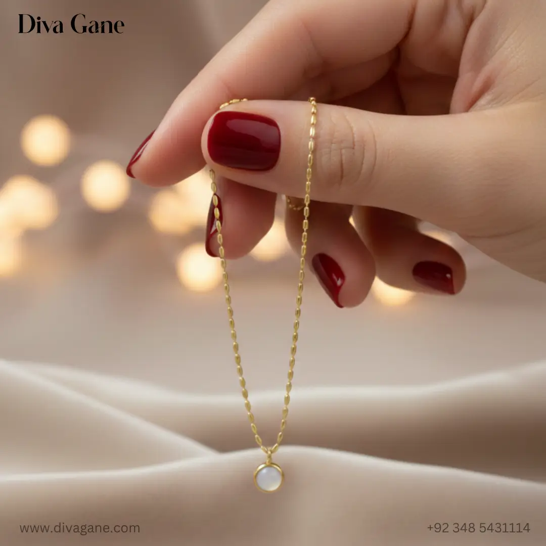 the luna minimalist moonstone pendant Close-up of a hand with deep red nail polish holding a dainty gold necklace with a circular white stone pendant against a soft bokeh background