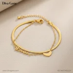 About Double-layered gold bracelet featuring a thick flat snake chain and a thin link chain with 'Lover' script and heart charm.