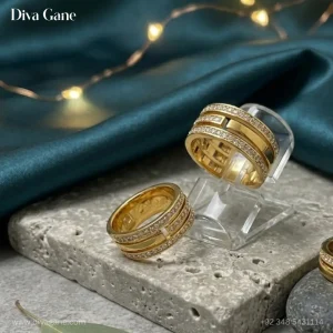 A wide 18K gold plated triple-band ring with pavé crystal rows resting on a gray stone pedestal with green velvet and fairy lights