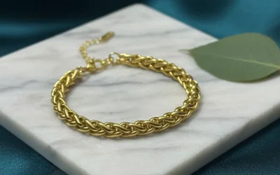 Top-down view of a gold woven link bracelet placed on a white marble square with a green leaf accent and bokeh fairy lights